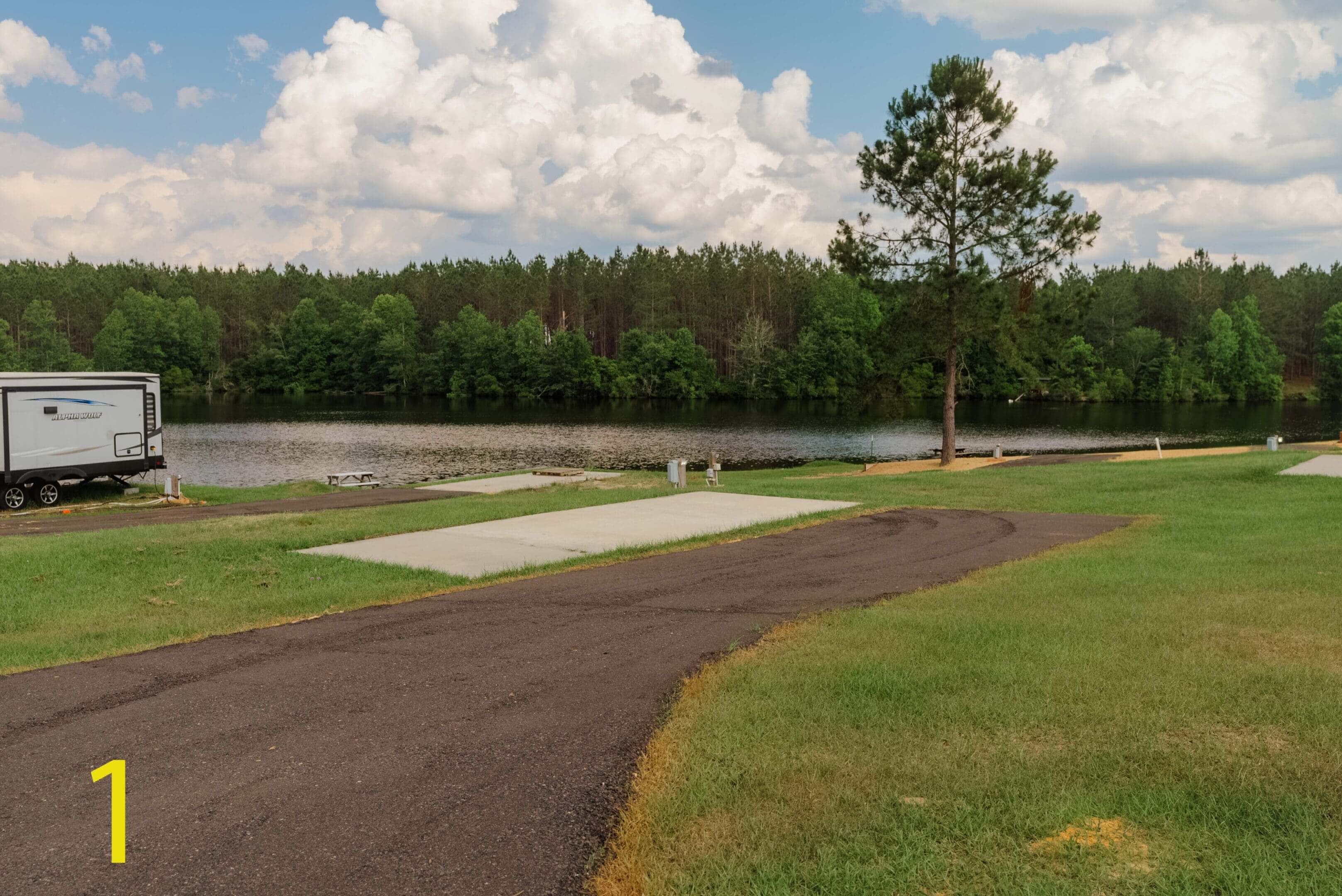 A winding path beside a calm pond with trees in the background under a cloudy sky.